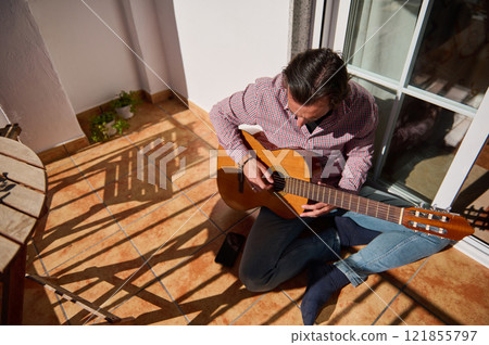 Man playing an acoustic guitar while sitting on a sunny terrace 121855797