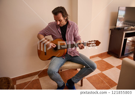 Man playing a guitar indoors seated on a light-colored chair Man playing a guitar indoors seated on a light-colored chair 121855803