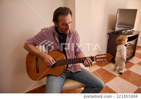 Father playing acoustic guitar while child walks in a cozy living room 121855804