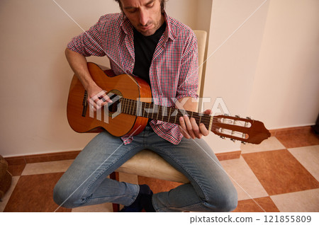 Man playing acoustic guitar indoors wearing casual clothes sitting on chair 121855809