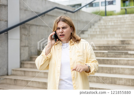 Angry woman on cell phone walking outside building near steps. Frustrated woman talking on phone. Summer time. Negative people emotion Angry woman on cell phone walking outside building near steps. Frustrated woman talking on phone. Summer time. Negative people emotion 121855880
