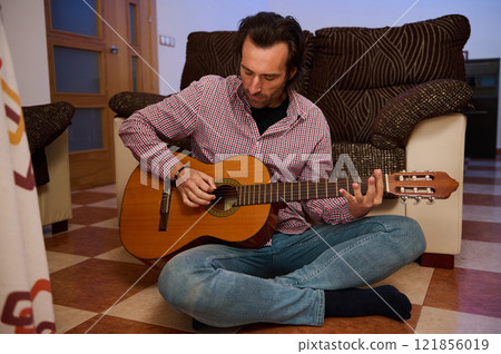 Man playing acoustic guitar while sitting cross-legged on the floor Man playing acoustic guitar while sitting cross-legged on the floor 121856019