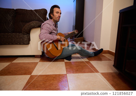 Man playing acoustic guitar while sitting on tiled living room floor Man playing acoustic guitar while sitting on tiled living room floor 121856027