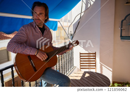 Man playing an acoustic guitar on a sunny balcony outdoors Man playing an acoustic guitar on a sunny balcony outdoors 121856062