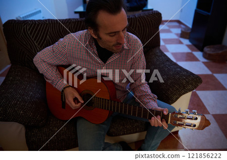 Man playing a guitar while sitting on a couch in a cozy living room 121856222