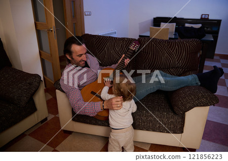 Father playing guitar with young child in warm family living room setting 121856223