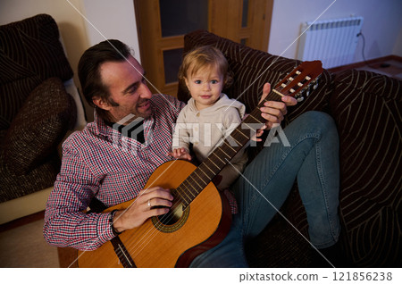 Father and child playing acoustic guitar together indoors on the couch 121856238
