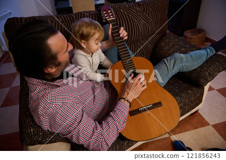 Father showing toddler how to play guitar while sitting at home 121856243