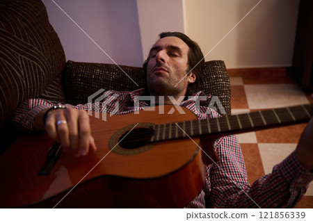 Man relaxing with a guitar lying on a cozy sofa indoors Man relaxing with a guitar lying on a cozy sofa indoors 121856339