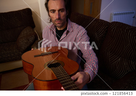 Man inspecting an acoustic guitar while sitting indoors on a cozy couch 121856413