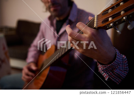 Person playing acoustic guitar at home showcasing focus and skilled fingerwork 121856447