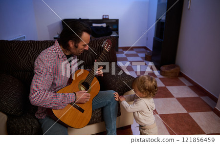 Man playing guitar while toddler watches in a home environment Man playing guitar while toddler watches in a home environment 121856459