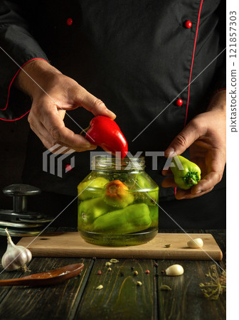 A chef skillfully adds vibrant red and green peppers into a jar filled with herbs and spices, creating a flavorful stuffed pepper dish. The rustic kitchen ambiance enhances the culinary artistry 121857303