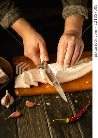 Hands skillfully slice through fresh pork on a wooden cutting board, surrounded by garlic cloves and a bright red chili pepper. A serene kitchen atmosphere enhances the culinary craft 121857306