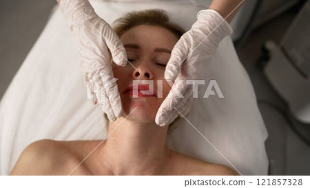 Wiping face with cotton pad. Close-up portrait of cosmetologist's hands cleaning woman's face with cotton pads. Cosmetologist wiping client's face on couch with cotton pads. Wiping face with cotton pad. Close-up portrait of cosmetologist's hands cleaning woman's face with cotton pads. Cosmetologist wiping client's face on couch with cotton pads. 121857328