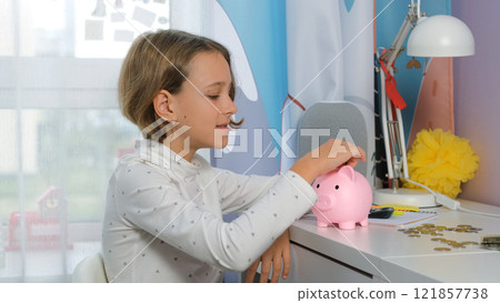 In a bright cozy room, a young girl places coins into a pink piggy bank, learning about personal finance and the importance of saving money. Personal finances and managing money 121857738
