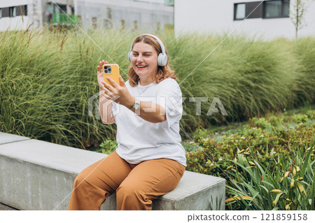 Portrait of young happy woman walking on the street in the city. Girl talking on a video call with phone and headphones outdoor, blogger records stories. Online communication concept. 121859158