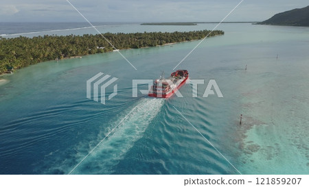 Red cargo ship sails in clear turquoise water of Maupiti, French Polynesia, leaving white wake, with lush greenery and mountain in background. Export and import. Wild travel destination, remote nature Red cargo ship sails in clear turquoise water of Maupiti, French Polynesia, leaving white wake, with lush greenery and mountain in background. Export and import. Wild travel destination, remote nature 121859207
