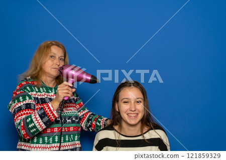 Caucasian woman in her 40s blow-drying her teenage daughter's hair, isolated on blue background. 121859329