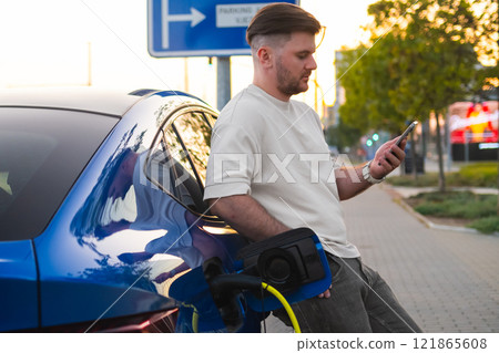Man charges his electric vehicle while using a mobile phone in the street at sunset. Man charges his electric vehicle while using a mobile phone in the street at sunset. 121865608