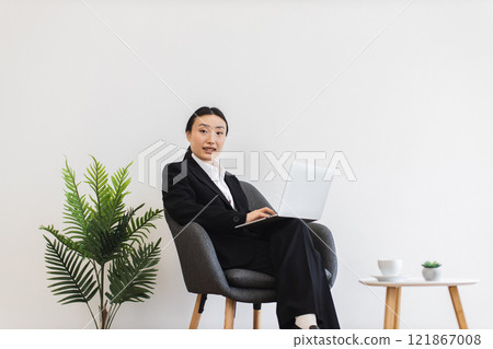 Asian businesswoman sitting on chair using laptop in modern office. Young adult female wearing business suit. Minimalist workspace design with natural plant decor suggests professional environment. 121867008