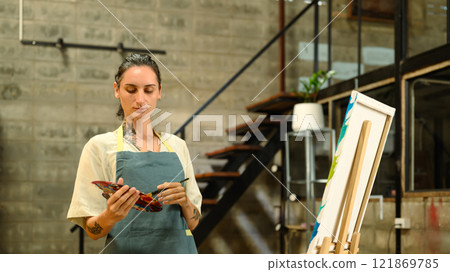 Focused Caucasian artist in an apron holding paint palette working on her art in the industrial style studio Focused Caucasian artist in an apron holding paint palette working on her art in the industrial style studio 121869785