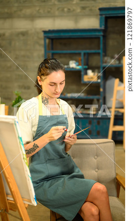 Caucasian artist in an apron sitting near an easel preparing their paint palette in her studio 121869797