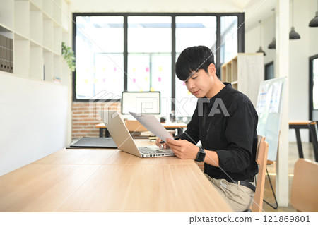 Young entrepreneur in a black shirt reviewing papers while working on his laptop in modern office Young entrepreneur in a black shirt reviewing papers while working on his laptop in modern office 121869801