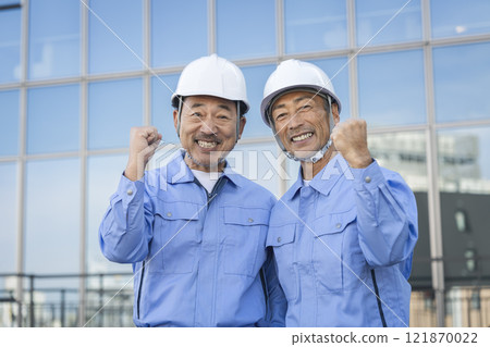 Two middle-aged men in work clothes making a fist pump while looking at the camera Two middle-aged men in work clothes making a fist pump while looking at the camera 121870022