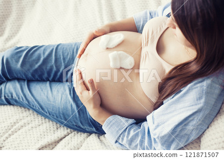 A pregnant woman is seen resting comfortably on bed, holding a pair of baby shoes near her belly. She appears content and serene, enjoying a quiet moment A pregnant woman is seen resting comfortably on bed, holding a pair of baby shoes near her belly. She appears content and serene, enjoying a quiet moment 121871507