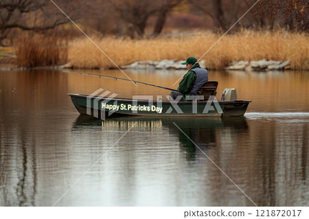 A man is fishing on a small boat with "Happy St. Patricks Day" written on it, in a calm lake. Concept of peaceful holiday fishing. 121872017