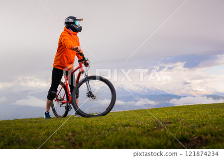 man rides a mountain bike on top of a green hill. A guy in a protective helmet stands on a meadow looking into the distance, with large snow-capped mountains in the background among clouds 121874234
