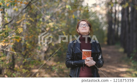 Smiling city woman relaxing in the forest. Thoughtful woman in glasses and office clothes walking in the autumn park, breathing fresh air, enjoying nature. 121875359