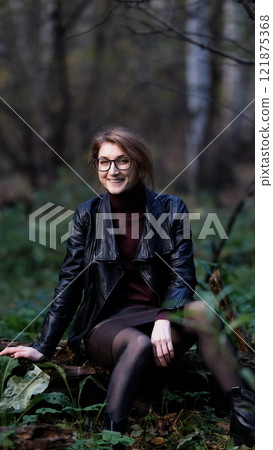 Smiling city woman relaxing in the forest. Thoughtful woman in glasses and office clothes sitting casually on an old fallen tree covered with moss outdoors, breathing fresh air, enjoying nature. 121875368