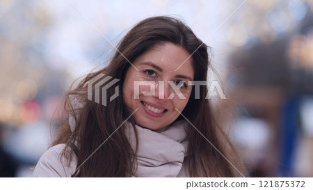 Beautiful woman with long brown hair, in warm winter clothes, standing on a snowy street in winter. She smiles and enjoys the snow, laughs and looks at the camera. Close-up 121875372