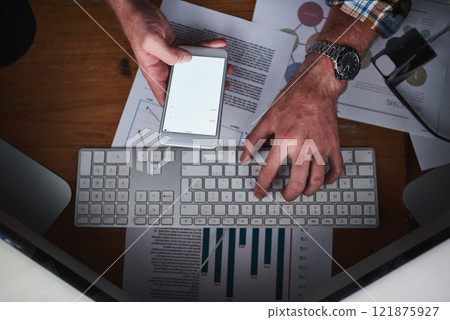 Hands on a computer keyboard and typing on a phone in an office with paper work from above. Closeup of a business man browsing the internet, finishing a report or sending a communication email Hands on a computer keyboard and typing on a phone in an office with paper work from above. Closeup of a business man browsing the internet, finishing a report or sending a communication email 121875927