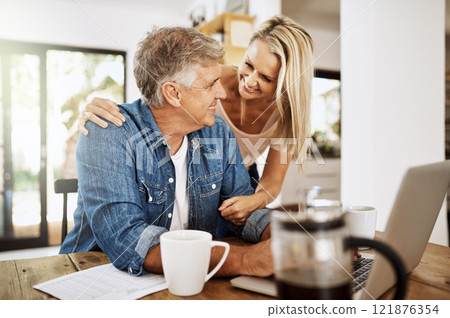 Mature couple talking and browsing on a laptop while spending time together. Loving and happy wife hugging and distracting her freelancing husband while working from home at the kitchen table Mature couple talking and browsing on a laptop while spending time together. Loving and happy wife hugging and distracting her freelancing husband while working from home at the kitchen table 121876354