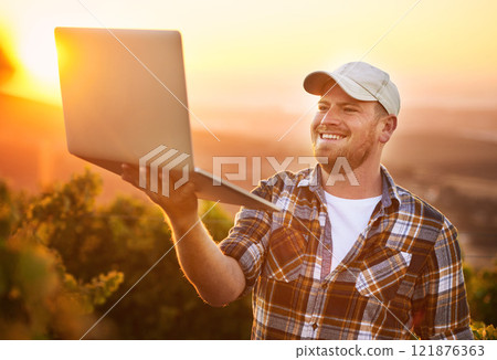A casual, happy and smiling young male farmer holding a laptop in a vineyard with flare. A relaxed, carefree and natural outdoors man using technology to make working easier on a hot summer day A casual, happy and smiling young male farmer holding a laptop in a vineyard with flare. A relaxed, carefree and natural outdoors man using technology to make working easier on a hot summer day 121876363