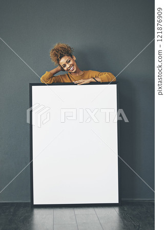 A beautiful confident woman holding a blank whiteboard or billboard sign with copy space. Happy and attractive female with a smile standing behind a banner. Young lady advertising with a poster 121876909