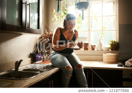 Browsing social media on tablet, searching online and reading funny stories, gossip and articles on technology app in home kitchen. Smiling and happy woman sitting on countertop and playing games 121877088