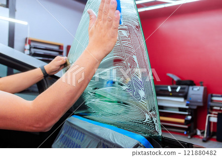 Worker is carefully applying window tint film to car rear window in a well lit workshop setting. Worker is carefully applying window tint film to car rear window in a well lit workshop setting. 121880482