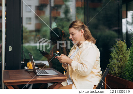 Smiling woman sitting in cafeteria holding smartphone and working on laptop. Successful businessman working in cafe outside. Young beautiful stylish woman with cup of coffee sitting in restaurant Smiling woman sitting in cafeteria holding smartphone and working on laptop. Successful businessman working in cafe outside. Young beautiful stylish woman with cup of coffee sitting in restaurant 121880929