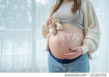 Young pregnant woman holding small knitted baby shoes on her belly near window at home in third trimester, with copy space. Concept of motherhood and bond between a mother and her unborn child Young pregnant woman holding small knitted baby shoes on her belly near window at home in third trimester, with copy space. Concept of motherhood and bond between a mother and her unborn child 121881509