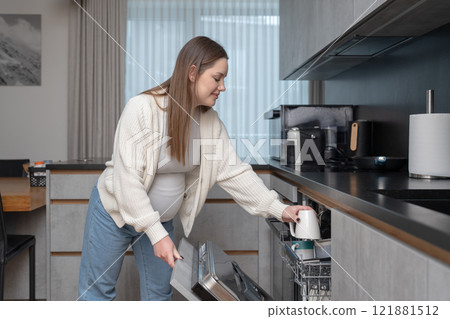 Young pregnant woman loading a dishwasher in the kitchen. Concept of daily life, home chores, and balancing household responsibilities during maternity. Young pregnant woman loading a dishwasher in the kitchen. Concept of daily life, home chores, and balancing household responsibilities during maternity. 121881512