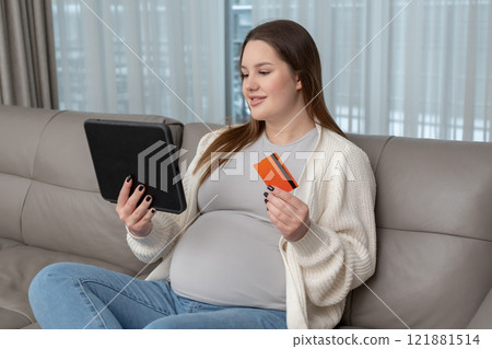 Pregnant woman using a tablet and credit card at home sitting on a sofa. Concept of online shopping, modern technology, digital transactions, and convenience during maternity preparation. 121881514
