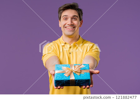 Man gives gift box by hands to camera on violet background. Guy smiling Man gives gift box by hands to camera on violet background. Guy smiling 121882477