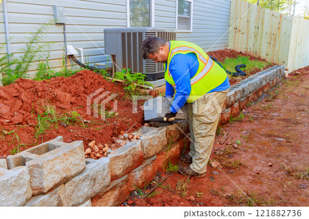 Construction worker carefully places stones to build sturdy retaining wall in home area. 121882736
