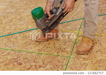 Carpenter worker operates air nail gun to fasten boards as part of flooring installation in construction project. 121882757