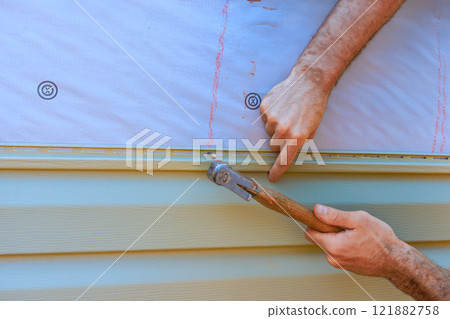 Construction worker skillfully secures siding board trim to exterior of house using tool, showcasing meticulous workmanship. Construction worker skillfully secures siding board trim to exterior of house using tool, showcasing meticulous workmanship. 121882758