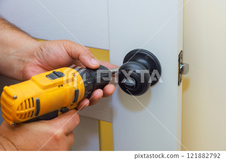 Carpenter is securing black door handle to white door with power drill, on task at hand. 121882792